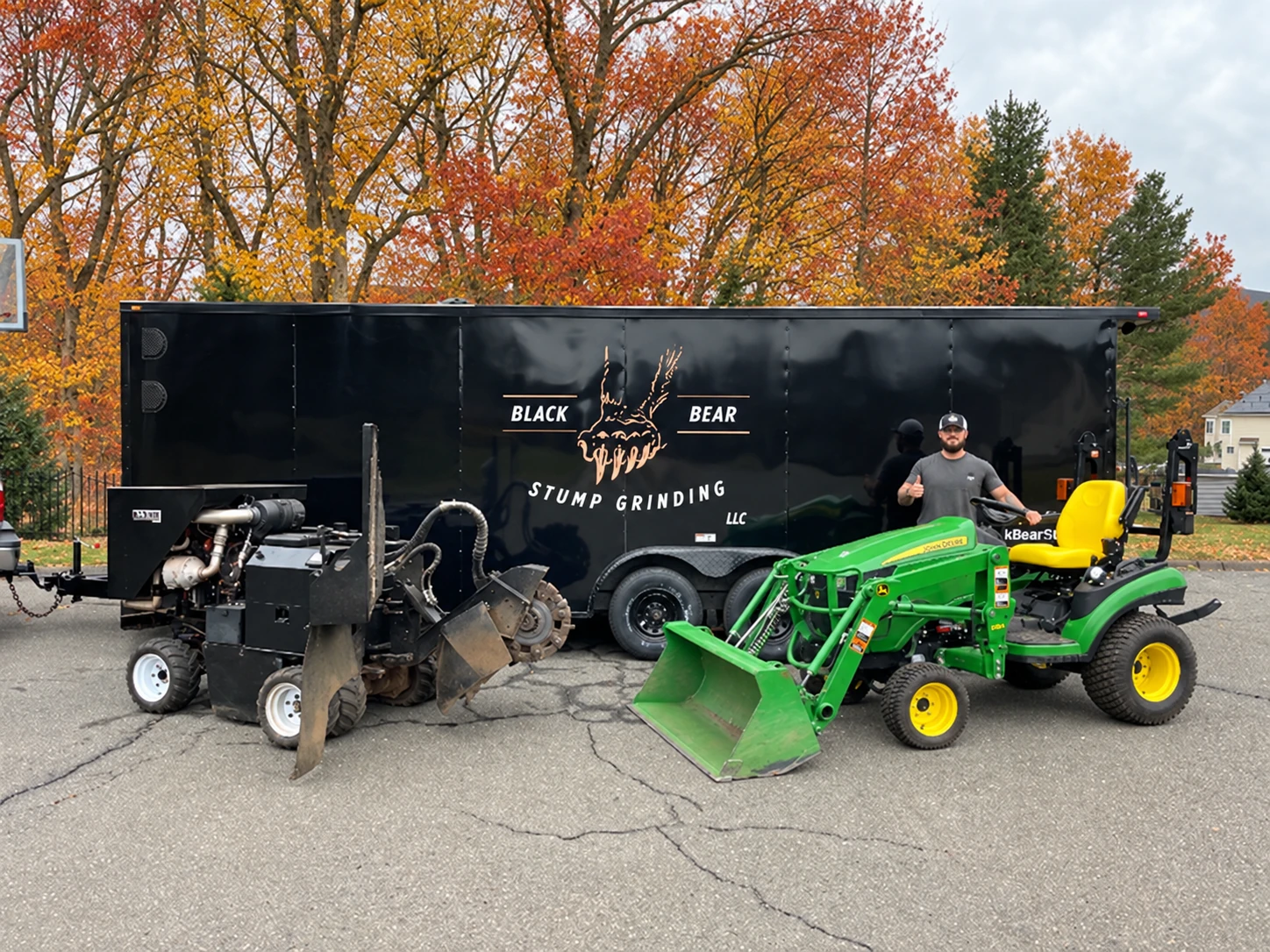 Alex beside the Black Bear Stump Grinding trailer, stump grinder, and tractor, with autumn trees behind